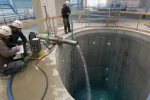 Tecnici con caschi osservano l’acqua che entra da una tubazione in una profonda vasca cilindrica in cemento