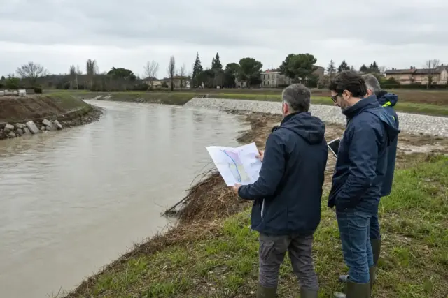 Tecnici in impermeabile controllano l’argine di un fiume in piena con una mappa e un tablet, cielo coperto