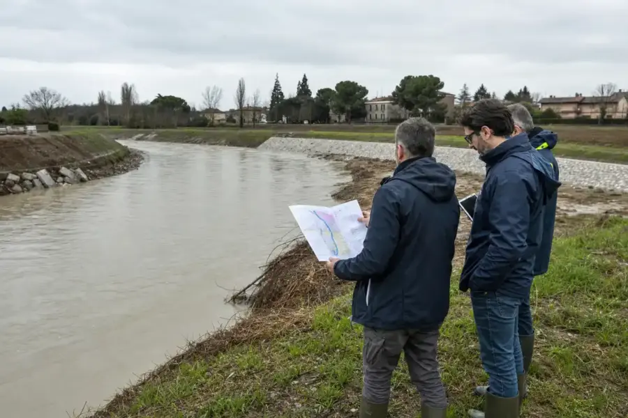 Tecnici in impermeabile controllano l’argine di un fiume in piena con una mappa e un tablet, cielo coperto