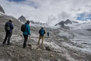 Escursionisti con casco e zaini osservano un ghiacciaio alpino in ritiro tra rocce e neve sporca, sotto un cielo nuvoloso