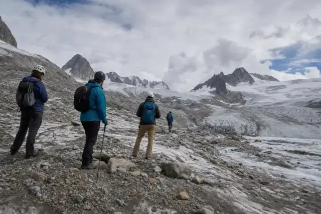 Escursionisti con casco e zaini osservano un ghiacciaio alpino in ritiro tra rocce e neve sporca, sotto un cielo nuvoloso