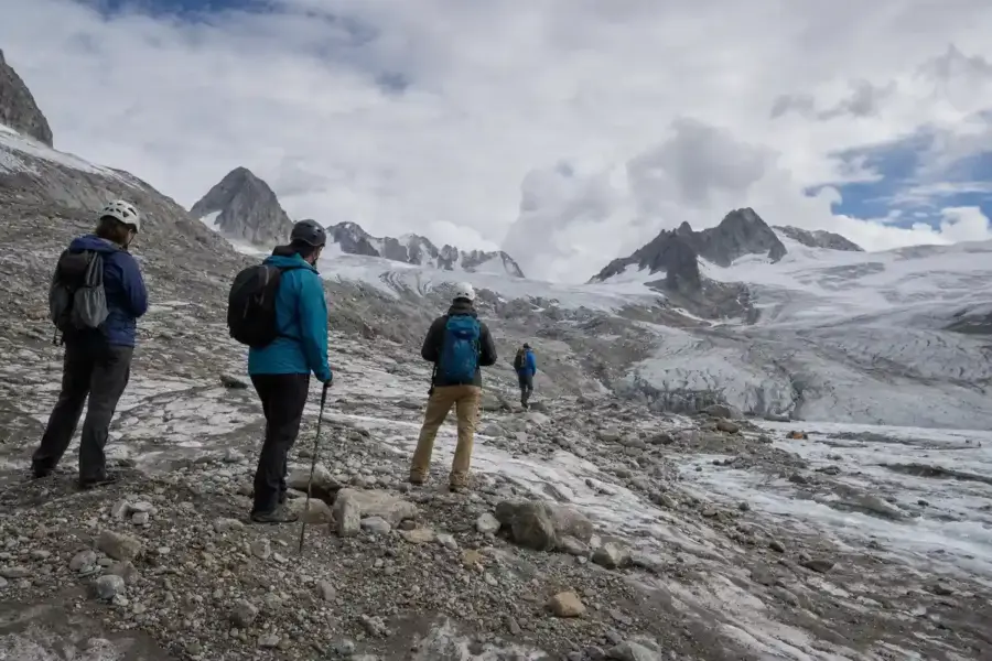 Escursionisti con casco e zaini osservano un ghiacciaio alpino in ritiro tra rocce e neve sporca, sotto un cielo nuvoloso