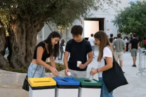 Tre giovani differenziano bicchieri e bottiglie in bidoni colorati accanto a un grande ulivo, in un cortile con pubblico