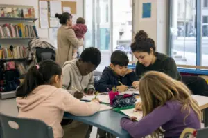 Bambini fanno compiti e disegni a un tavolo con un’educatrice in un centro di supporto con scaffali di libri