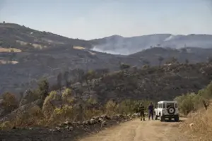 Colline bruciate dopo un incendio boschivo, con fumo in lontananza e due operatori vicino a un fuoristrada