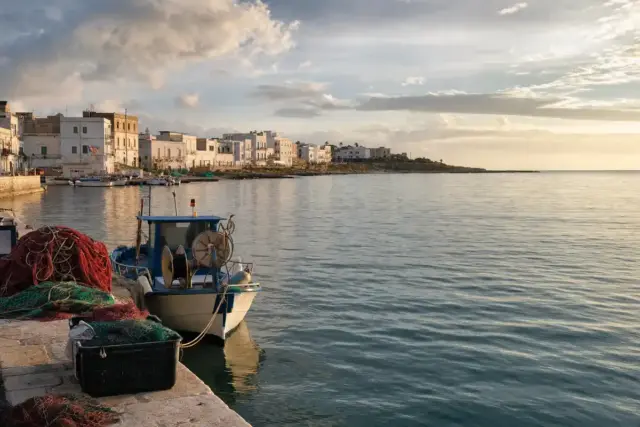 Barca da pesca ormeggiata in un porto costiero con reti sul molo e mare calmo sotto un cielo nuvoloso