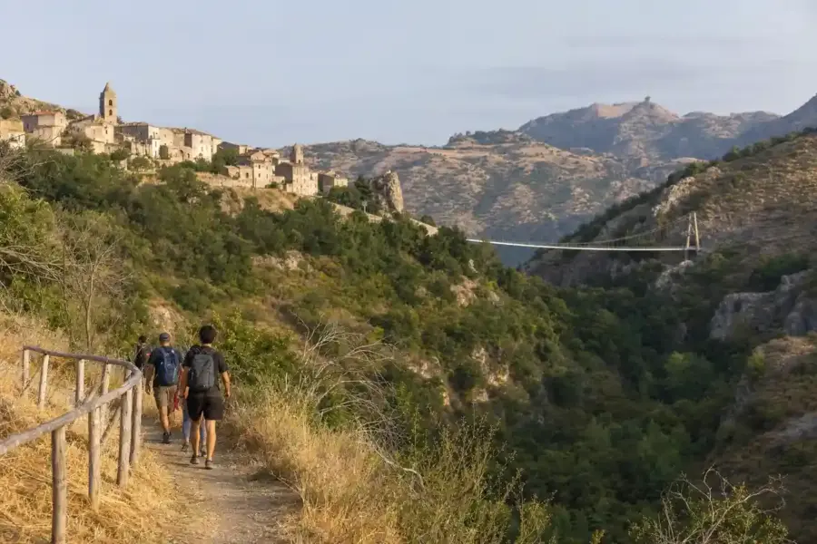 Escursionisti su un sentiero verso un borgo collinare e un ponte sospeso sopra una valle verde