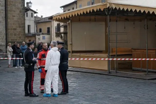 Carabinieri e soccorritore davanti a uno stand da festa transennato con nastro rosso e bianco in una piazza