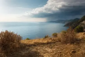 Costa mediterranea vista dall’alto con terra arida e crepata in primo piano e nuvole temporalesche all’orizzonte