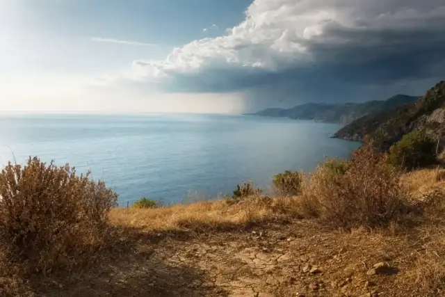 Costa mediterranea vista dall’alto con terra arida e crepata in primo piano e nuvole temporalesche all’orizzonte