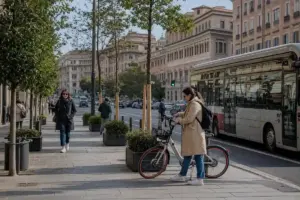 Pendolare con bici condivisa sul marciapiede alberato mentre passa un autobus in una strada urbana trafficata