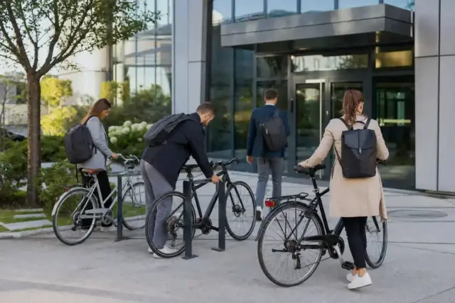 Dipendenti arrivano in bici davanti a un ingresso di uffici moderno, con biciclette al portabici e zaini